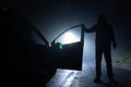 A man standing next to a car, with the door open, underneath a street light. On a foggy winters night