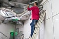A man standing on the stairs is repairing the ventilation ducts. Maintenance of communications is performed in the technical room