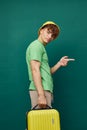 man stands on a background, dressed in a yellow hat, holding a bright travel suitcase in his hand, looking pleasantly Royalty Free Stock Photo