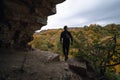 A man stands on the edge, at the exit from the cave. View of the autumn forest, tree crowns. Dramatic gray clouds. Travel and