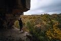 A man stands on the edge, at the exit from the cave. View of the autumn forest, tree crowns. Dramatic gray clouds. Travel and