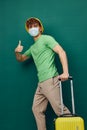 man stands on a green background, dressed in a yellow panama hat and with a mask on his face, holding a bright travel Royalty Free Stock Photo