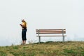 A man stands on a hilltop with a phone in his hands against a background of thick fog. An empty bench and a young man standing