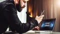 Man stands near table with laptop with inscription on monitor- cloud library, looks at screen of smartphone. Online education