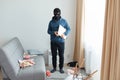 Man steals laptop in someone else house, runs away, being of persecution, wears robber mask and blue hoodie, standing near sofa Royalty Free Stock Photo