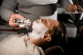 Man stylist applying shaving foam onto client& x27;s face using old-fashioned shaving brush in barbershop Royalty Free Stock Photo