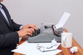 The man in a suit typing on an old-fashioned typewriter. On the table are glasses and there is a mug. Neutral background