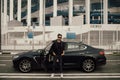 A man in sunglasses and a leather jacket stands near a sports car