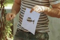 Man in t-shirt and denim pants holds in his hand sheet of white paper with fish stencil and demonstrates in park.