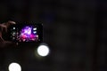 Man takes photo with his phone of crowd against backdrop of music scene on street in night city.