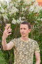 man takes selfie on his cell phone on white lilacs blooming background, close-up, side view Royalty Free Stock Photo