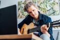 Man taking online guitar lessons with a laptop at home sitting on the gray couch, sofa in the living room. He watching Royalty Free Stock Photo