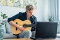 Man taking online guitar lessons with a laptop at home sitting on the gray couch, sofa in the living room. He watching Royalty Free Stock Photo