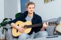 Man taking online guitar lessons via a mobile application on his phone at home sitting on the gray couch, sofa in the Royalty Free Stock Photo