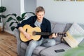 Man taking online guitar lessons via a mobile application on his phone at home sitting on the gray couch, sofa in the Royalty Free Stock Photo