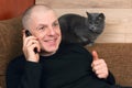 Man talking on the phone resting on the couch next to a gray cat