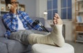 Man talking on the phone while sitting on the sofa at home, with his injured foot resting on a chair Royalty Free Stock Photo