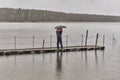 Man talking on phone and walking under umbrella on wooden pier on rainy gray day over of backdrop of lake and forest.