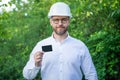 Man taskmaster in hardhat showing blank contact card outdoors, copy space