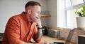 Man, thinking and typing on laptop at home, planning and reflection by kitchen window in apartment. Male person Royalty Free Stock Photo