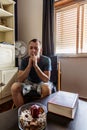 A man with a thoughtful look sits in a chair opposite the window in the room. On the table in front of him is a large red book and Royalty Free Stock Photo