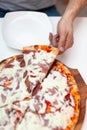 Man took first slice of delicious Italian pizza. In frame hand taking slice of hot pizza with ham on wooden board Royalty Free Stock Photo