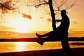 Man on tree. Silhouette of lone man sit on branch of birch tree at sunset at shoreline. Royalty Free Stock Photo