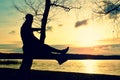 Man on tree. Silhouette of lone man sit on branch of birch tree at sunset at shoreline. Royalty Free Stock Photo