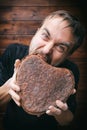 Man try greedily bites off piece of rye rough bread in shape of heart, concept of greed and hunger, brown background