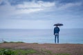 A man with an umbrella in the rain stands on a cliff and looks at the ocean back view. tourist place.
