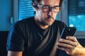 Man using cell phone at home office in low key interior. Casual male typing text message on smartphone while sitting at the desk