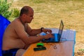 Man using his laptop sitting at folding picnic table in a camping. Working while traveling. Freelance work. Work and travel Royalty Free Stock Photo