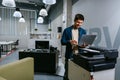 Man using laptop in modern office environment while standing near printer in bright work space Royalty Free Stock Photo