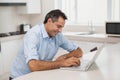 Mature man sitting at kitchen counter using silver laptop, wearing light blue shirt, natural light Royalty Free Stock Photo