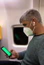 Man using smart phone with green screen wearing a face mask and earphones in airplane seat Royalty Free Stock Photo