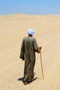 A man walking alone in the middle of the desert wearing a galabya and holding a stick