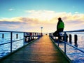 Man in warm jacket and baseball cap sit on pier and enjoy quiet morning sea. Tourist relax. Royalty Free Stock Photo