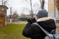 Man in warm jacket and cap with back pack standing back to camera and taking photographs of abandoned building