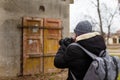 Man in warm jacket and cap with back pack standing back to camera and taking photographs of abandoned building