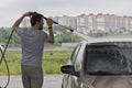 A man washes a car at a car wash, watering car with hose from a tap. City service for washing cars. A young man with a beard