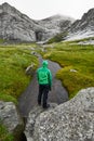 Young man with a green jacket and hood is standing with his back on a rock in front of a huge mountain on Lofoten Islands in Norwa Royalty Free Stock Photo