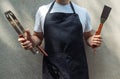 A man wearing black chef`s apron, holding barbecue tools: bbq tongs, spatula