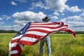 Man wearing cowboy hat standing outdoors in beautiful grass field, holding American flag blowing in wind behind him, blue sky