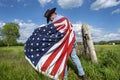 Man wearing cowboy hat walking in grass field, near barbwire fence, American flag wrapped around his shoulders