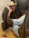 Man wearing cowboy straw hat and boots sitting down pondering, feet on the table