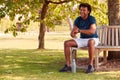 Man Wearing Fitness Clothing Sitting On Seat Under Tree Checking Activity Monitor On Smartwatch