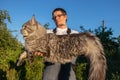 A man wearing glasses and overalls is holding a huge, gray Maine Coon cat