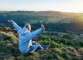 Man wearing grey coat and jeans raising his hands high and sitting on top of the mountain Royalty Free Stock Photo