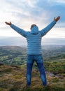 Man wearing grey coat and jeans raising his hands high and standing on top of the mountain Royalty Free Stock Photo