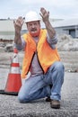 Man wearing hard hat and safety vest kneeling next to traffic cone gesturing with his hands Royalty Free Stock Photo